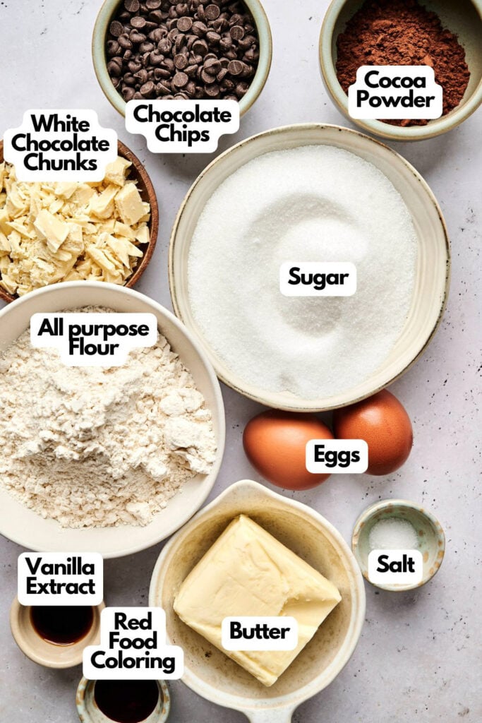 Bowls and plates contain labeled baking ingredients for red velvet brownies: white chocolate chunks, chocolate chips, cocoa powder, sugar, all-purpose flour, eggs, vanilla extract, red food coloring, butter, and salt.