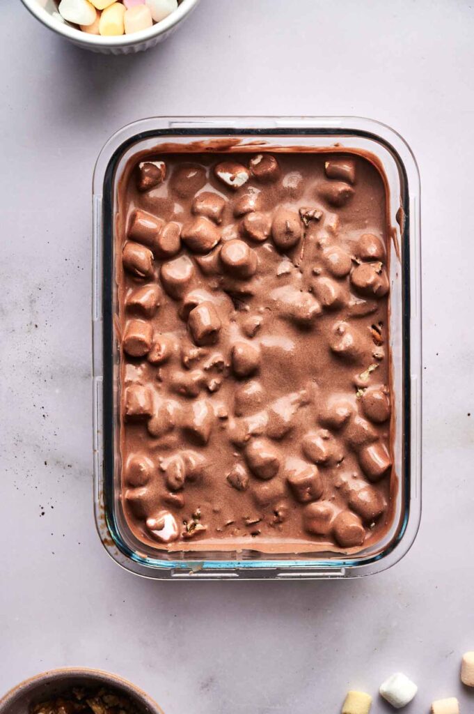 A rectangular glass container filled with rocky road ice cream&mdash;chocolate ice cream mixed with mini marshmallows&mdash;sits on a light surface.