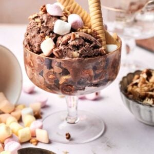 Rocky road ice cream in a glass bowl topped with mini marshmallows, wafer sticks, and nuts, with spoons and extra toppings on the table.