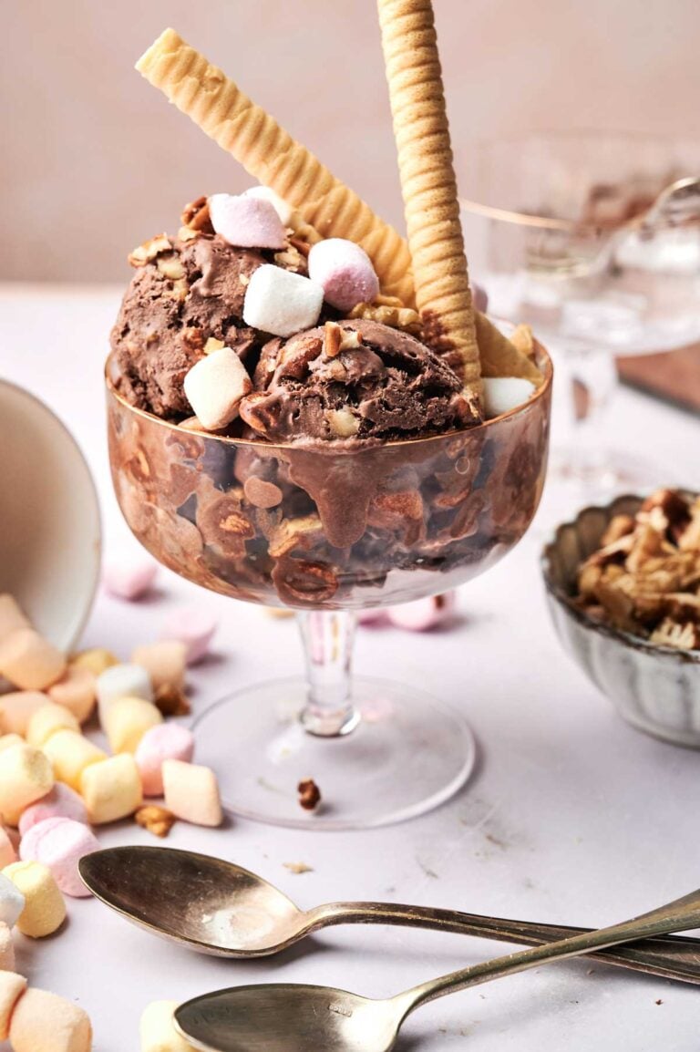 Rocky road ice cream in a glass bowl topped with mini marshmallows, wafer sticks, and nuts, with spoons and extra toppings on the table.