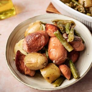 A bowl of roasted potatoes and green beans, inspired by Sausage Green Bean Potato Casserole, is seasoned with herbs and sits on a light-colored surface next to a bottle of olive oil and a white casserole dish.