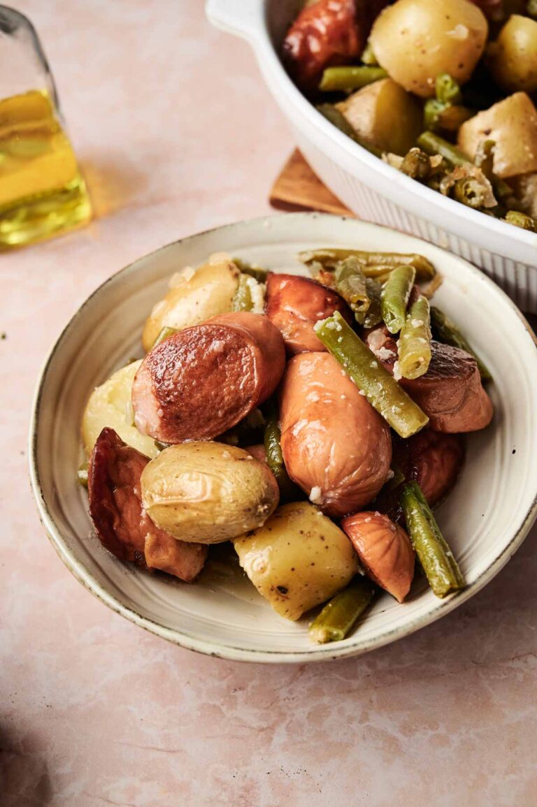 A bowl of roasted potatoes and green beans, inspired by Sausage Green Bean Potato Casserole, is seasoned with herbs and sits on a light-colored surface next to a bottle of olive oil and a white casserole dish.