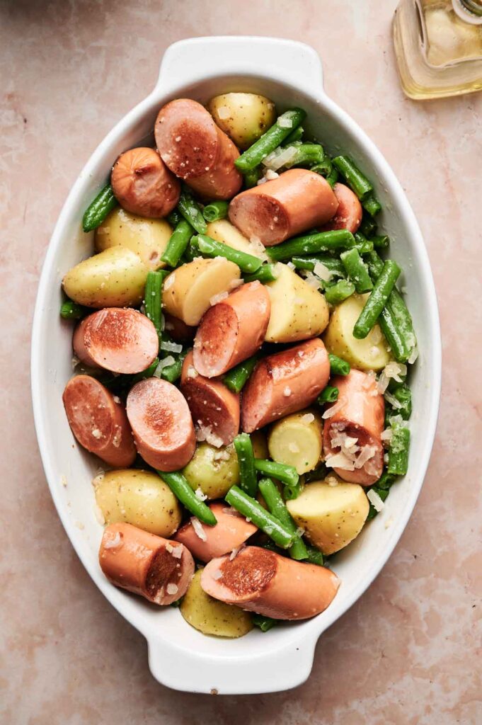 An oval dish filled with sliced sausages, halved baby potatoes, green beans, and chopped onions—perfect for serving a hearty Sausage Green Bean Potato Casserole.