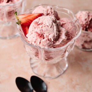 A glass dish filled with Strawberry Ice Cream, each scoop garnished with a fresh strawberry; two black spoons and a strawberry slice rest nearby.