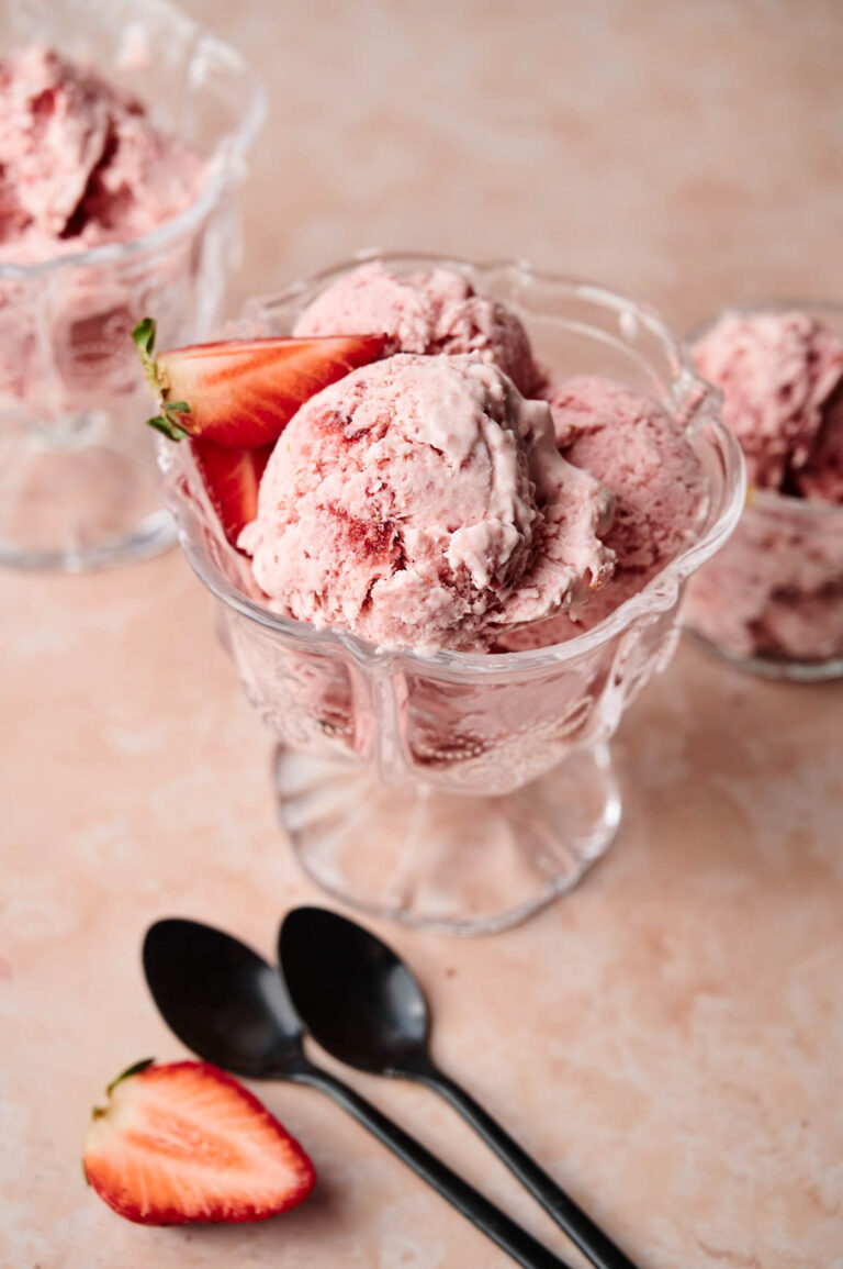 A glass dish filled with Strawberry Ice Cream, each scoop garnished with a fresh strawberry; two black spoons and a strawberry slice rest nearby.