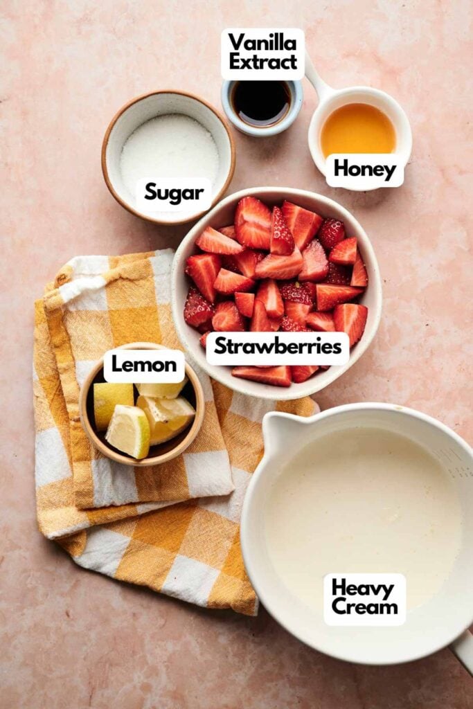 Bowls and cups containing ingredients for strawberry ice cream&mdash;strawberries, heavy cream, lemon wedges, sugar, vanilla extract, and honey&mdash;are arranged on a peach-colored surface with a yellow checked cloth.