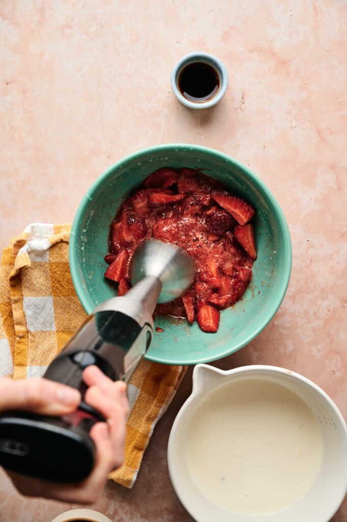A hand uses an immersion blender to puree strawberries for strawberry ice cream in a teal bowl, with cream, vanilla, and a yellow towel nearby on a pink surface.