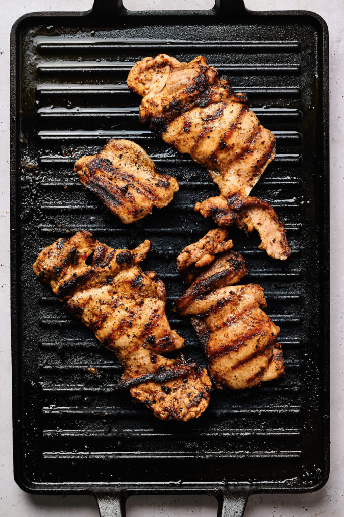 Four pieces of grilled, seasoned chicken cook on a black grill pan with distinct char marks&mdash;perfect for adding to a savory Street Corn Chicken Rice Bowl.