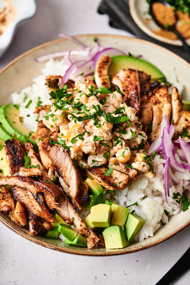 A Street Corn Chicken Rice Bowl featuring white rice topped with grilled chicken strips, avocado slices, red onion, chickpeas, and chopped herbs.