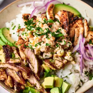 A Street Corn Chicken Rice Bowl featuring white rice topped with grilled chicken strips, avocado slices, red onion, chickpeas, and chopped herbs.