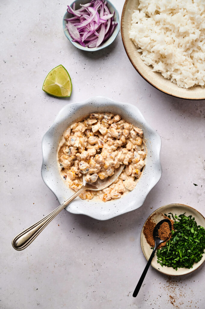 A bowl of creamy shrimp curry with a spoon, surrounded by white rice, sliced red onions, a lime wedge, chopped herbs, and spices&mdash;perfect alongside a Street Corn Chicken Rice Bowl for a flavorful meal on a light surface.