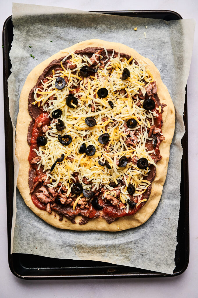 A homemade taco pizza topped with shredded cheese, sliced black olives, and pieces of meat on a parchment-lined baking sheet.