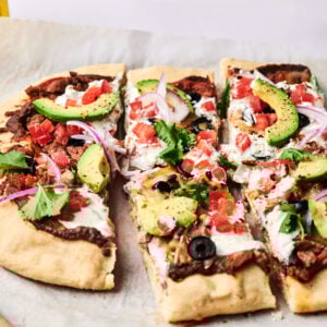 Rectangular taco pizza topped with avocado slices, tomato, black olives, red onion, cilantro, and white sauce, sliced and served on parchment paper over a wooden board.