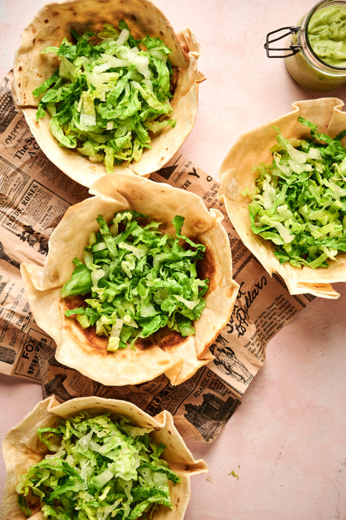 Five taco salad tortilla bowls filled with shredded lettuce and other fresh fillings are arranged on vintage-style paper, with a jar of green sauce nearby.
