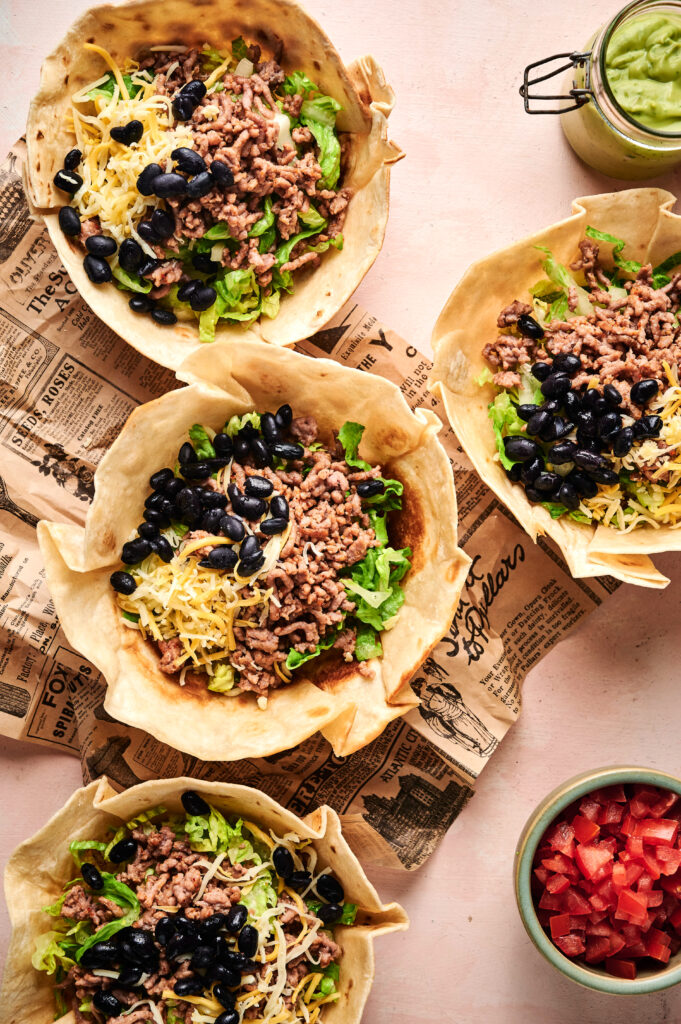 Four tortilla bowls filled with ground meat, black beans, lettuce, and shredded cheese create a delicious taco salad display on newspaper-print paper, with sides of diced tomatoes and guacamole nearby.