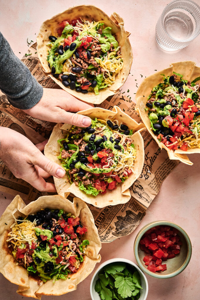 Four tortilla bowls filled with taco salad ingredients, including lettuce, cheese, beans, tomatoes, and beef. A hand holds one bowl. Nearby are bowls of chopped tomatoes and cilantro.