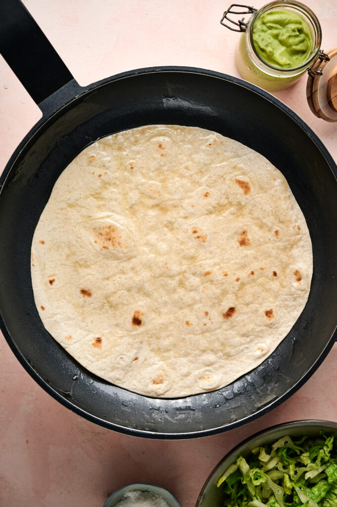 A flour tortilla is being heated in a black skillet for a Taco Salad. Nearby are jars of guacamole, shredded lettuce, and other ingredients on a pink surface.