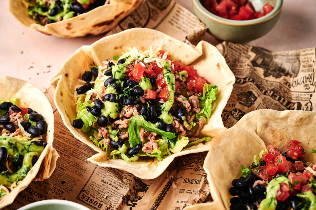A taco salad in a crispy tortilla bowl is filled with lettuce, ground meat, black beans, guacamole, shredded cheese, and diced tomatoes, all served atop a sheet of newspaper.