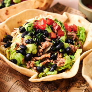 A classic Taco Salad served in an edible tortilla bowl, topped with ground meat, black beans, shredded lettuce, tomatoes, and diced avocado on newspaper print paper.