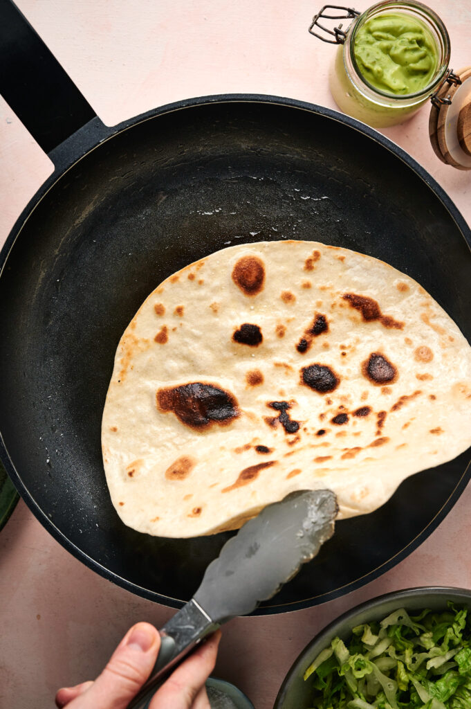 A hand holds tongs, flipping a piece of flatbread in a black skillet; bowls of green sauce and chopped lettuce, perfect toppings for a Taco Salad, are nearby.