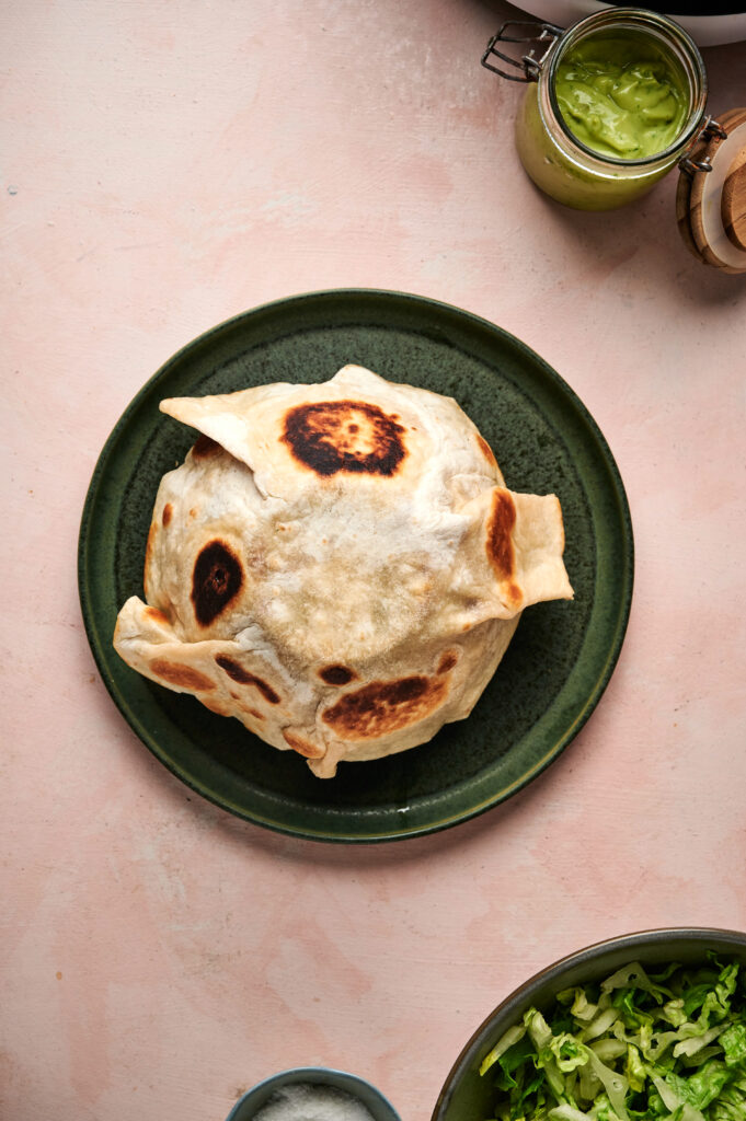 A round, puffed flatbread with charred spots sits on a green plate; jars of sauce and a bowl of lettuce&mdash;perfect fixings for a fresh taco salad&mdash;are partially visible nearby.