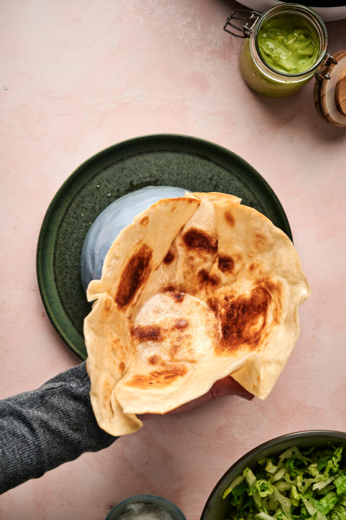 A hand holds a piece of browned flatbread over a green plate, surrounded by jars and bowls filled with sauces and greens, resembling the vibrant fixings of a taco salad, all arranged on a light pink surface.