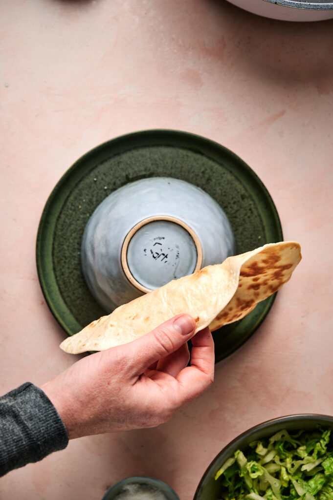A hand holds a folded tortilla over an upside-down bowl placed on a green plate, preparing the perfect base for a Taco Salad, with some chopped lettuce visible in the corner.