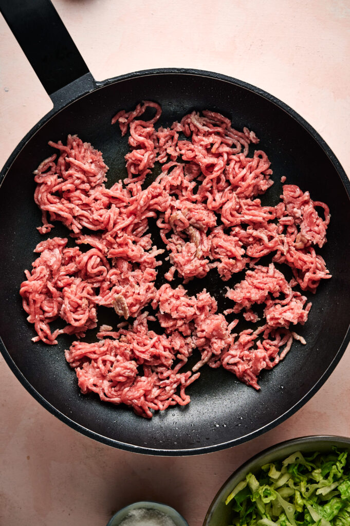 Raw ground beef spread out in a black frying pan, ready to be cooked for a delicious taco salad. A small bowl of salt and a bowl of chopped lettuce are visible nearby.