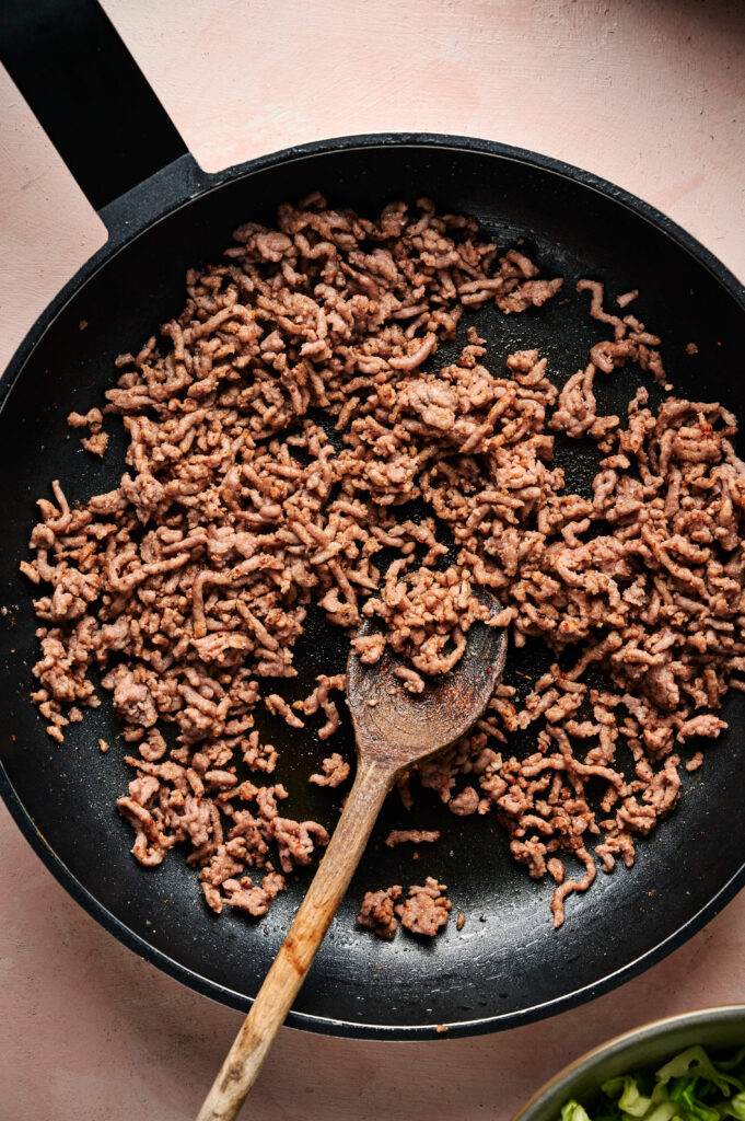 Cooked ground beef in a black skillet with a wooden spoon, perfect for Taco Salad, viewed from above on a light pink surface.