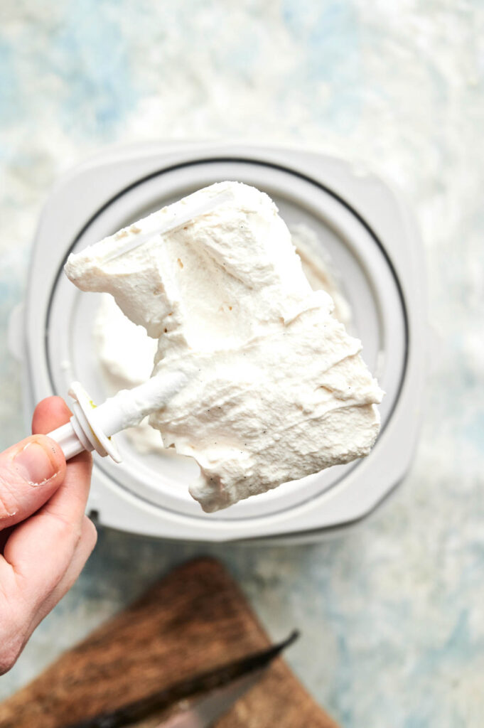 A hand holds a mixer attachment covered in thick, freshly whipped cream above a mixing bowl on a light surface, ready to be folded into homemade vanilla ice cream.