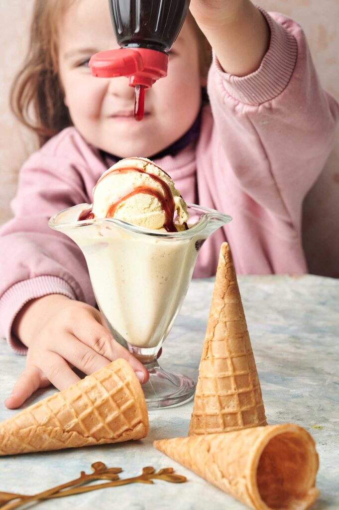 A child pours chocolate syrup onto a glass of creamy vanilla ice cream, with empty waffle cones lying on the table in front.