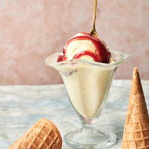 A glass dish of Vanilla Ice Cream with red syrup and a decorative spoon sits beside two empty waffle cones on a marble surface.