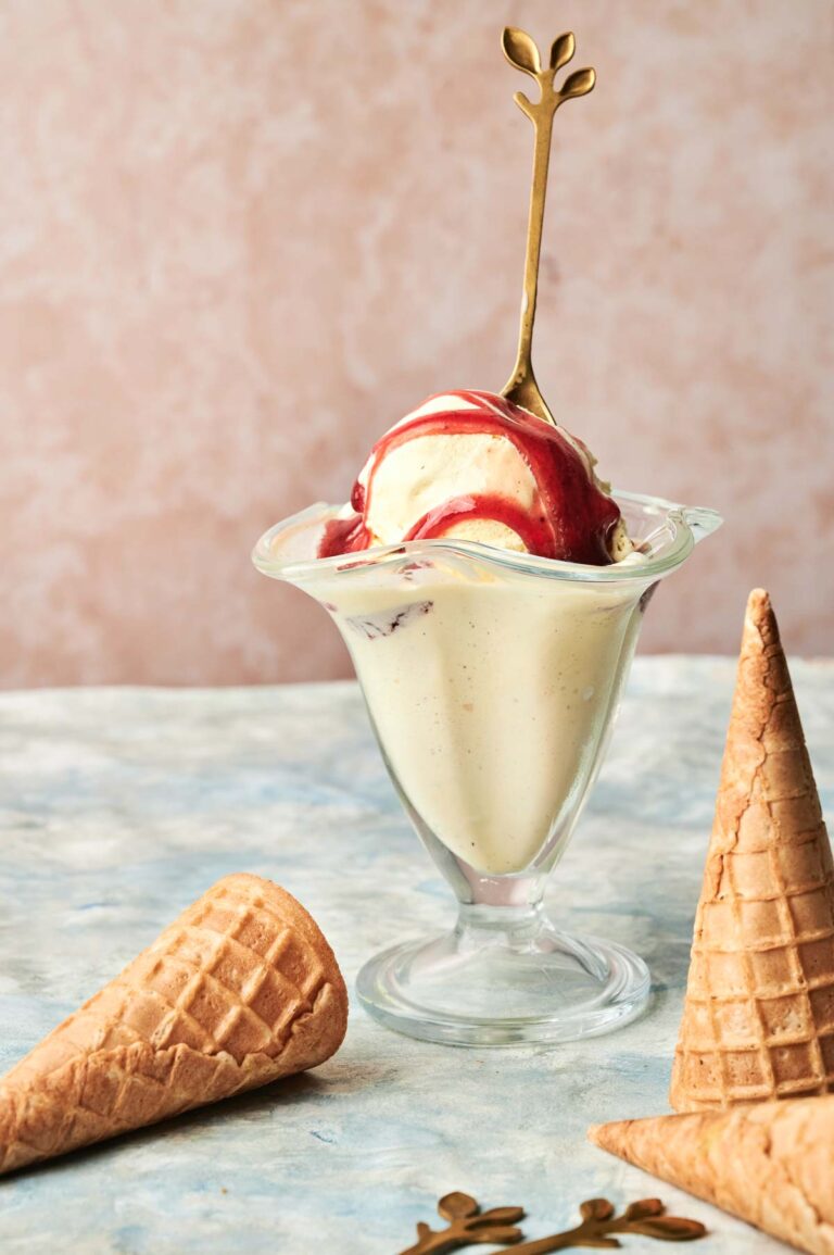 A glass dish of Vanilla Ice Cream with red syrup and a decorative spoon sits beside two empty waffle cones on a marble surface.