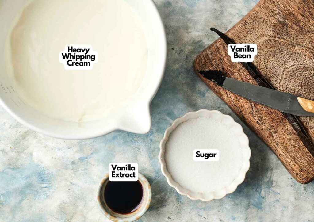 A bowl of heavy whipping cream, sugar, vanilla extract, and a vanilla bean sit on a cutting board with a knife&mdash;all labeled and ready for homemade Vanilla Ice Cream.
