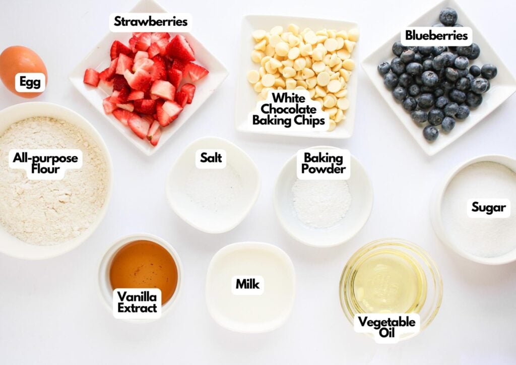 Top-down view of ingredients for 4th of July Muffins in bowls and plates, including flour, strawberries, blueberries, white chocolate chips, egg, salt, baking powder, sugar, vanilla extract, milk, and vegetable oil.
