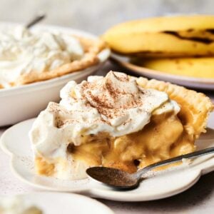 A slice of Banana Cream Pie topped with whipped cream and cinnamon sits on a white plate, with a spoon and fresh bananas in the background.