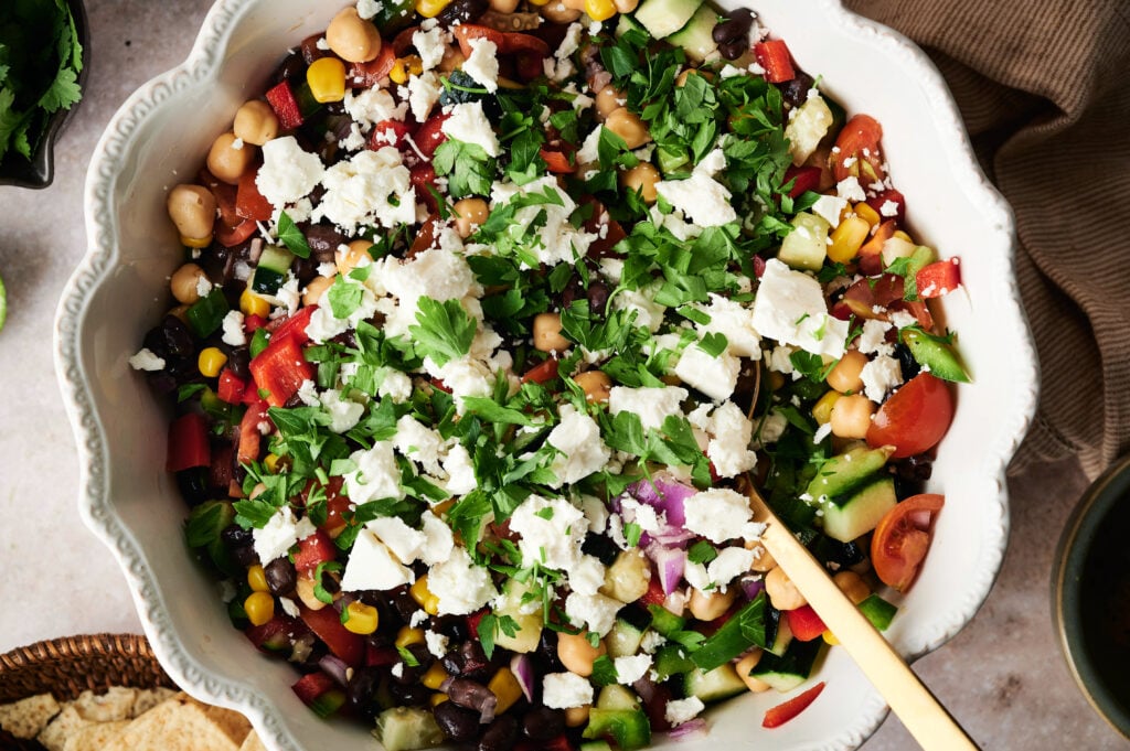 A bowl of Cowboy Caviar, featuring chickpeas, black beans, corn, tomatoes, cucumber, red onion, parsley, and crumbled feta cheese.
