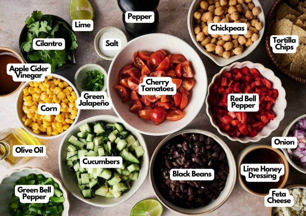 A variety of labeled ingredients in bowls on a table, including cherry tomatoes, black beans, corn, cucumbers, peppers, chickpeas, onion, feta, chips, and assorted seasonings—perfect for assembling your own cowboy caviar.