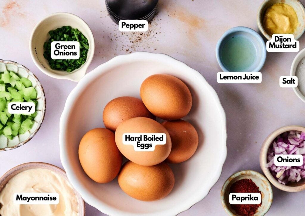 A bowl of hard boiled eggs, ready to be transformed into a classic egg salad, is surrounded by small dishes of mayonnaise, celery, green onions, Dijon mustard, lemon juice, paprika, pepper, salt, and onions.