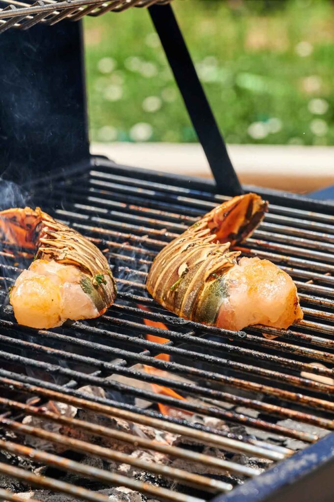 Two Grilled Lobster Tails sizzle over an open flame on a barbecue, smoke rising as they cook to perfection against a blurred outdoor backdrop.