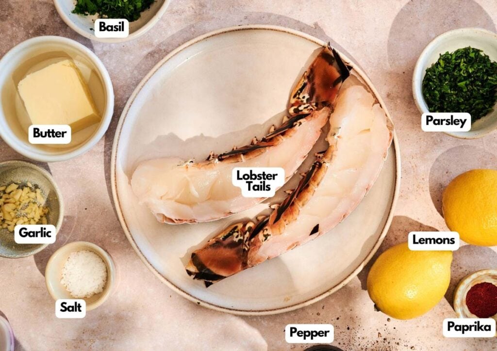 A plate with two raw lobster tails, perfect for Grilled Lobster Tail, surrounded by bowls of butter, chopped basil, parsley, minced garlic, salt, pepper, paprika, and two whole lemons.