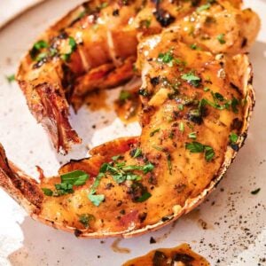 Two Grilled Lobster Tail halves rest on a plate, topped with creamy, seasoned sauce and garnished with chopped fresh herbs.