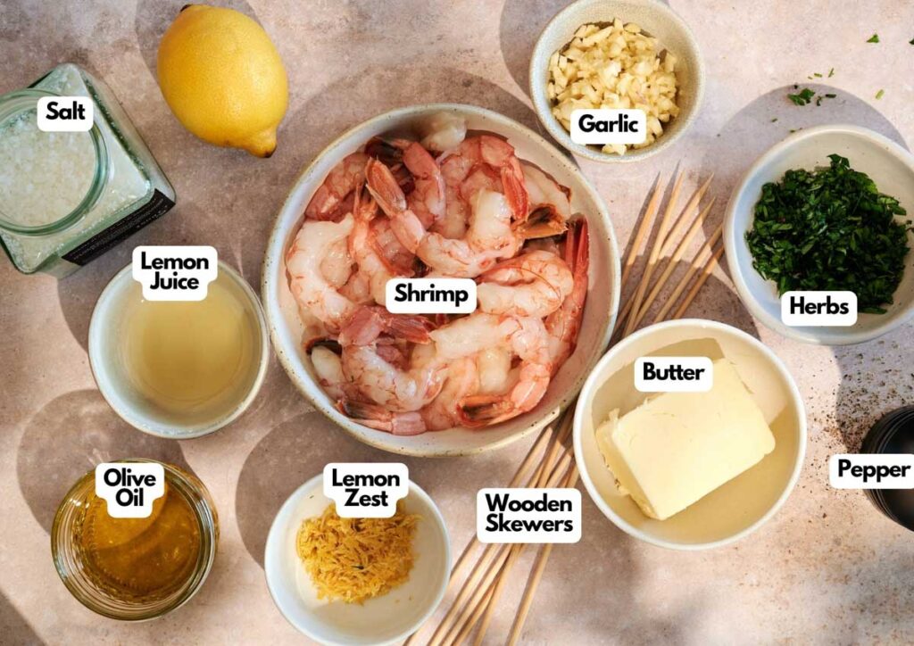 A bowl of shrimp ready for grilled shrimp sits surrounded by labeled bowls of salt, lemon juice, lemon zest, olive oil, garlic, chopped herbs, butter, pepper, and wooden skewers on a countertop.