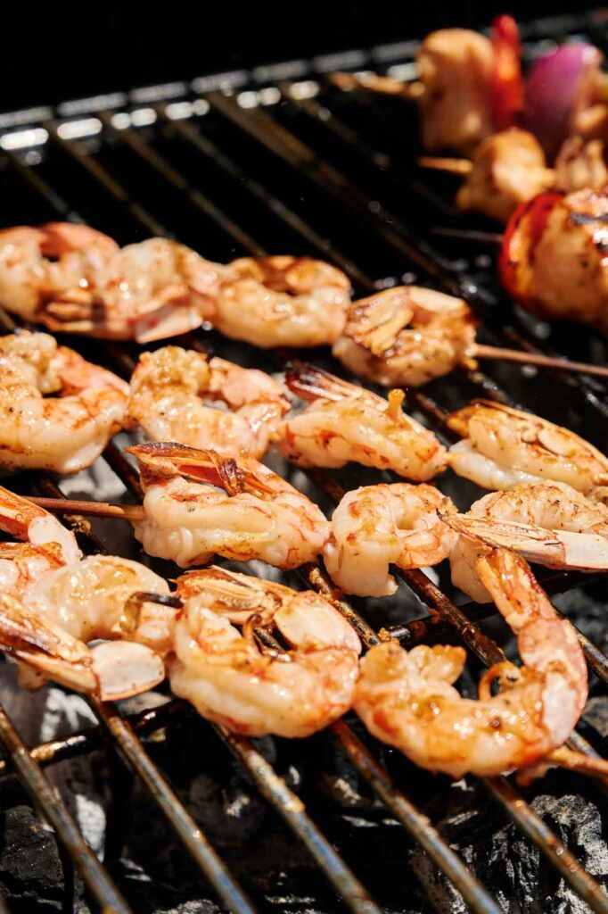 Grilled shrimp skewers are sizzling on the barbecue, with mushrooms and onions adding flavor in the background.