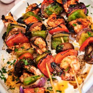 Grilled shrimp kabobs with bell peppers, onions, and mushrooms are served on a white plate, garnished with fresh herbs and sauce.