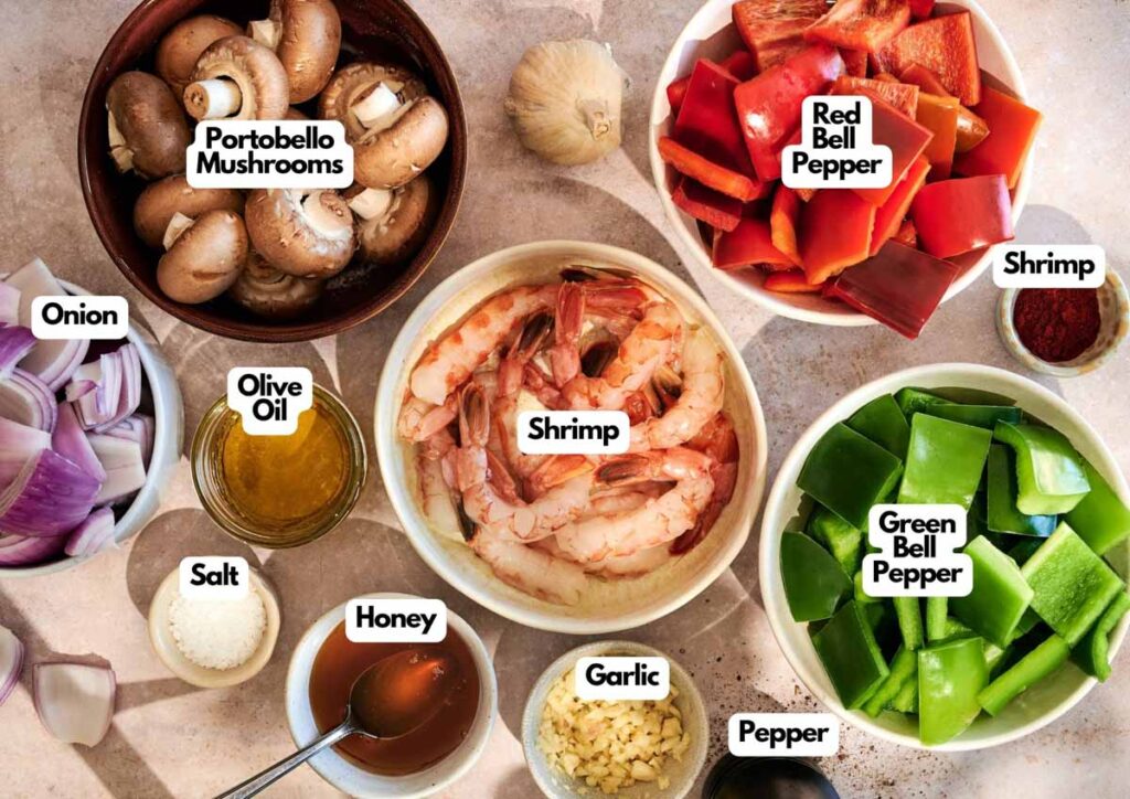 Various ingredients in bowls on a countertop, ready for grilled shrimp kabobs: raw shrimp, chopped onion, portobello mushrooms, red and green bell peppers, olive oil, salt, honey, garlic, and pepper.