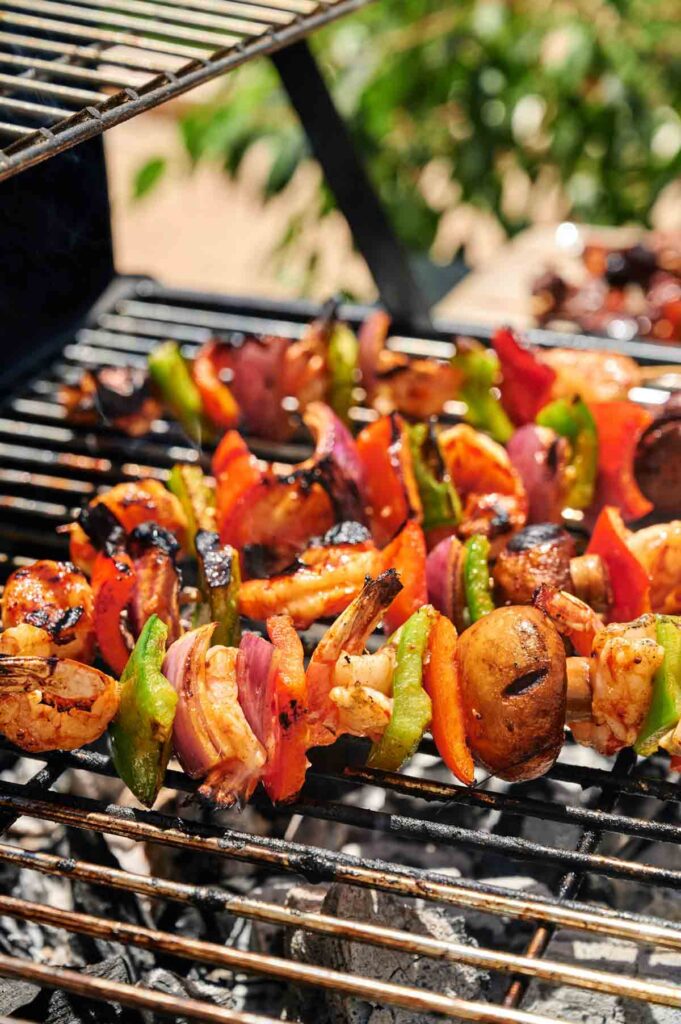 Colorful vegetable, chicken, and grilled shrimp kabobs sizzle over hot coals on an outdoor barbecue, displaying delicious char marks against a blurred background of lush greenery.