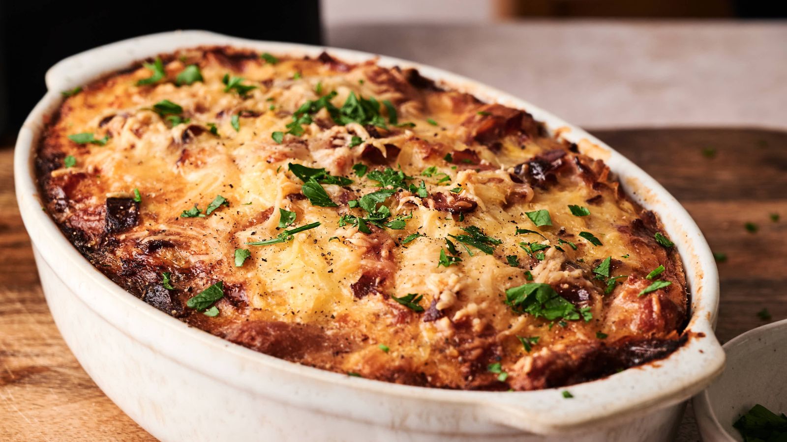 A baked casserole in an oval white dish, topped with melted cheese and chopped fresh herbs, sits on a wooden surface.