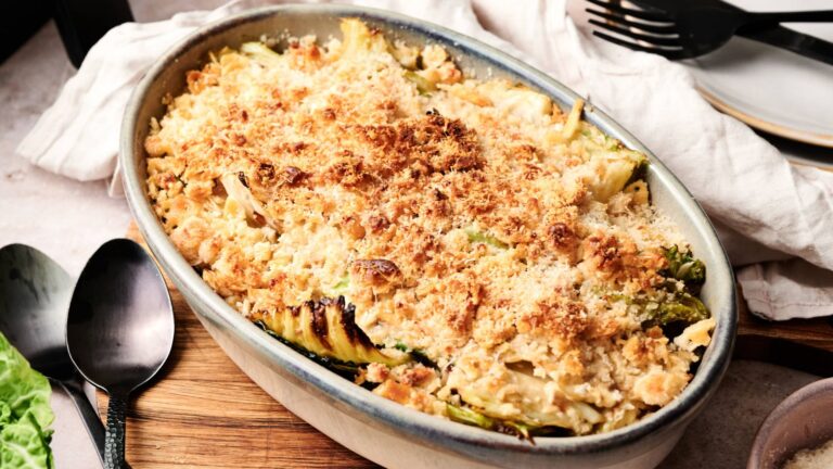 Oval ceramic dish filled with baked casserole topped with a golden breadcrumb crust, placed on a wooden board beside a spoon, fork, and napkin.