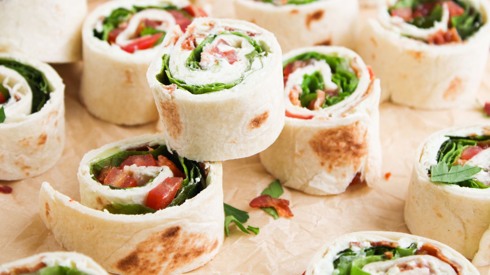 Several tortilla pinwheels filled with leafy greens, tomato, and possibly cheese or meat, arranged on parchment paper.